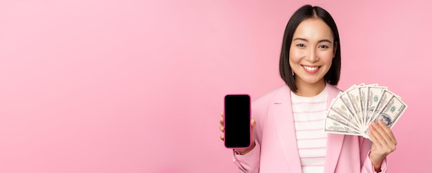 Image of korean successful corporate woman showing money dollars and smartphone app screen interface of mobile phone application concept of investment and finance pink background