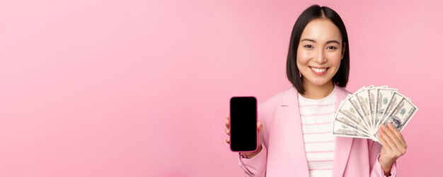 Image of korean successful corporate woman showing money dollars and smartphone app screen interface of mobile phone application concept of investment and finance pink background