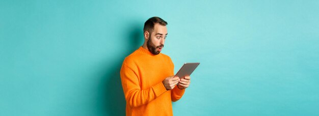 Immagine del modello maschile in maglione arancione che fissa lo schermo del tablet digitale che sembra sorpreso in piedi ov
