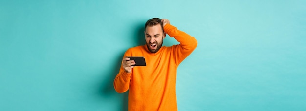 Free photo image of man looking confused at mobile phone watching something online standing puzzled against lig