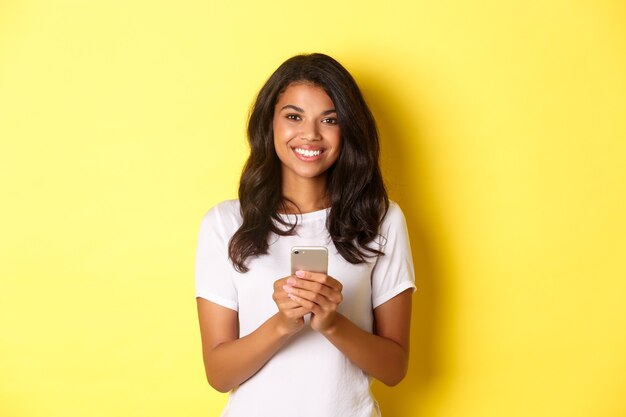 노란색 배경 위에 서 있는 휴대폰을 사용하여 웃고 있는 현대 아프리카계 미국인 소녀의 이미지