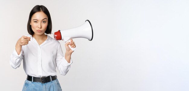 Image of modern asian woman with megaphone pointing at you camera making announcement white background