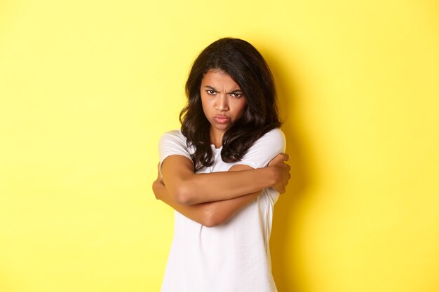 Image of moody cute african-american girl, hugging herself and sulking with offended expression, standing over yellow background.