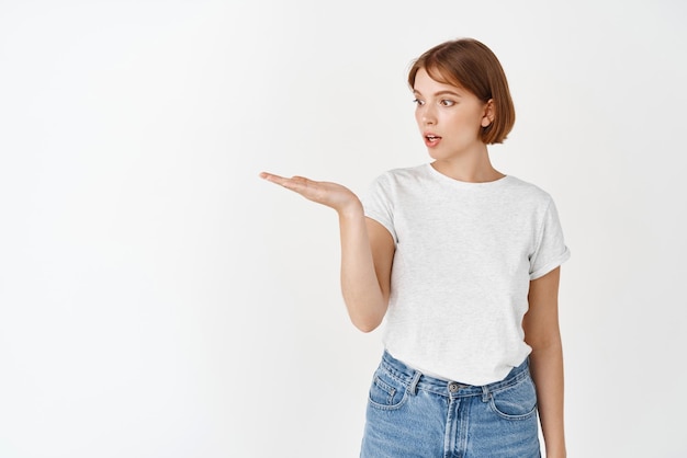 Image of natural young woman holding empty space on hand looking at palm excited and amazed standing on white background