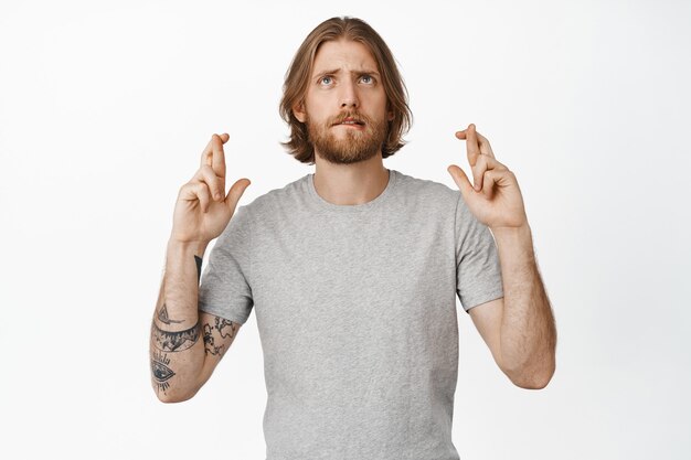 Image of nervous hopeful guy, blond man cross fingers and looking up with anxious face, wishing, waiting for dream come true, anticipating important news on white.