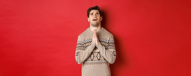 神に祈って、クリスマスの助けを懇願し、冬のセーターを着て、見上げて懇願し、赤い背景の上に立っている神経質で希望に満ちた男の画像。