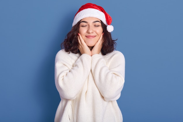 暖かい白い冬のセーターを着て素敵な若い女性のイメージとクリスマスのポーズを閉じた目と頬に手でポーズ、青い背景に分離されたポーズ、チャーミングでかわいい見えます。新しい耳のコンセプト。