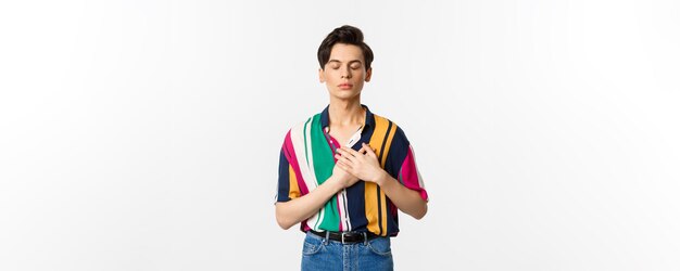 Image of nostalgic young gay man standing with closed eyes and hands on heart remember something sta