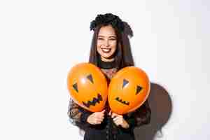 無料写真 怖い顔で2つのオレンジ色の風船を保持し、ハロウィーンを祝って、白い背景の上に立っている邪悪な魔女の衣装を着たアジアの女の子の画像。