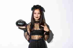 Бесплатное фото Изображение красивой азиатской женщины в готическом кружевном платье и венке указывая пальцем на черную тыкву, празднуя хэллоуин, нося костюм ведьмы, стоя над белой предпосылкой.