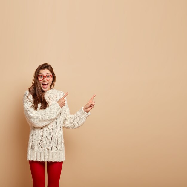 Бесплатное фото Изображение радостной, эмоциональной молодой девушки с довольным выражением лица, смеется от удовольствия, указывает в верхнем правом углу, рекламирует классный продукт, носит белый свитер, изолированное на бежевой стене