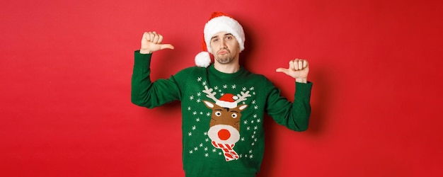 무료 사진 녹색 스웨터와 자신을 가리키는 산타 모자를 쓴 잘 생기고 자신감 있는 청년의 이미지
