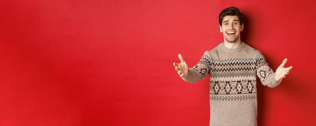 Изображение счастливого и польщенного красивого парня в рождественском свитере, протягивающего руки, чтобы поприветствовать гостей на новогодней вечеринке, приглашающего на празднование, стоящего на красном фоне Бесплатные Фотографии