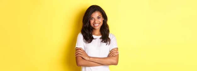 Бесплатное фото Изображение счастливой улыбающейся афроамериканской девушки в белой футболке, скрестившей руки на груди и выглядящей уверенно