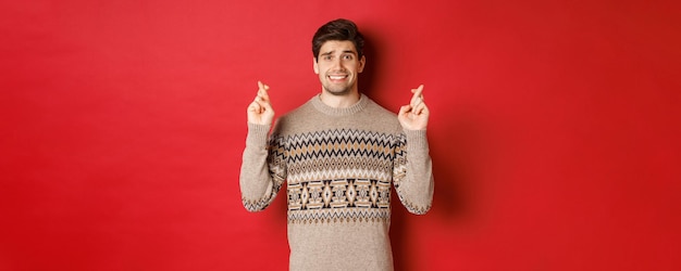 何かを待っている、幸運と願いを込めて指を交差させる、新年の贈り物、赤い背景に神経質なクリスマスセーターの希望と心配の男の画像 無料写真