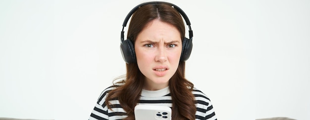 Бесплатное фото Изображение озадаченной молодой женщины в наушниках держит смартфон, хмурится и выглядит запутанной в камеру