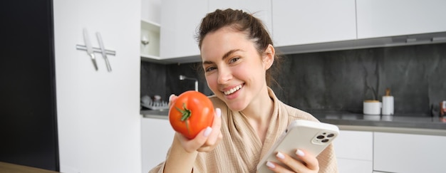 無料写真 キッチンに座る笑顔の美しい女性の画像がスマートフォンを持ち、トマトが注文に満足しているように見える