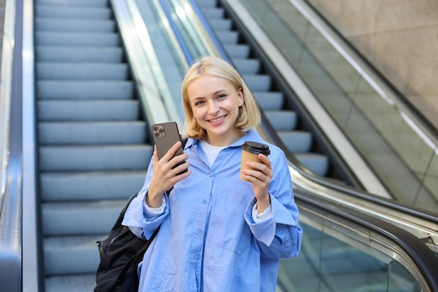 Бесплатное фото Изображение стильной молодой женщины в синей рубашке с чашечкой кофе и мобильным телефоном