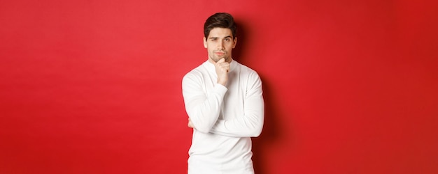 Изображение вдумчивого красивого человека, делающего предположение, думая и смотрящего в камеру, стоящего в белом свитере на красном фоне.