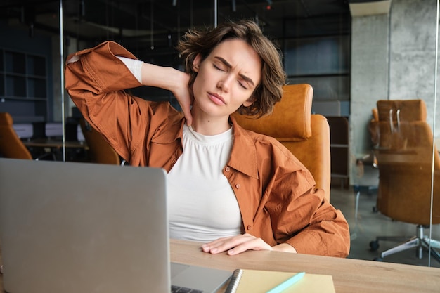 Бесплатное фото Изображение женщины с усталым лицом, сидящей с ноутбуком в офисе, чувствует напряжение в шее, боль в мышцах