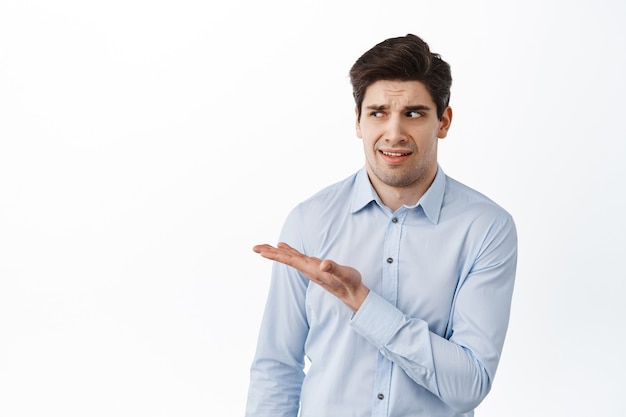 Image of office worker, businessman in shirt pointing, looking aside at something strange, standing confused and disappointed, white wall