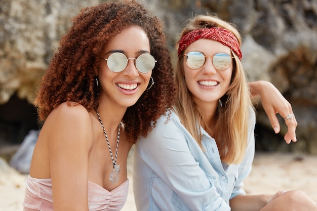 Free photo image of passionate homosexual couple embrace, wear trendy sunglasses. beautiful dark skinned young female hugs her female partner, looks at sunshine, sit together. happy moment. same sex relations