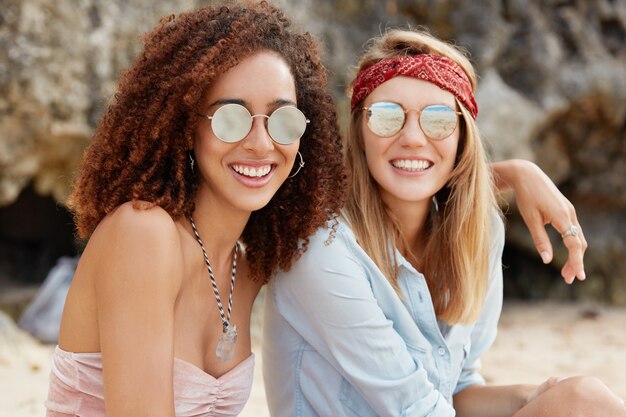 Image of passionate homosexual couple embrace, wear trendy sunglasses. Beautiful dark skinned young female hugs her female partner, looks at sunshine, sit together. Happy moment. Same sex relations