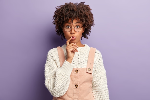 Image of pleasant looking young woman has folded lips, curly hair, keeps finger near mouth, listens attentively news, wears fashionable outfit, round spectacles, poses against purple wall