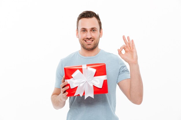 弓で赤いプレゼントボックスを押しながら笑顔とOKのサインを示す満足しているブルネットの男の画像