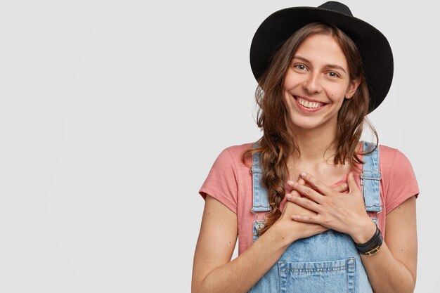 L'immagine del contadino sorridente e soddisfatto tiene le mani sul petto, esprime buon atteggiamento, ha i capelli scuri pettinati in una treccia, indossa copricapo e salopette, modelle contro il muro bianco con spazio libero a sinistra