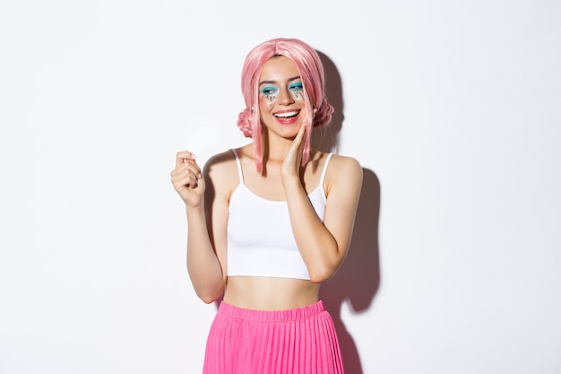 Image of pleased stylish girl in pink wig looking happy at credit card, dreaming of shopping, standing.