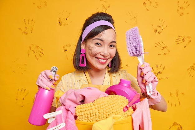 Image of positive housewife smiles pleasantly holds cleaning tools enjoys working process poses with brush and detergent poses near basket of laundry poses against yellow wall