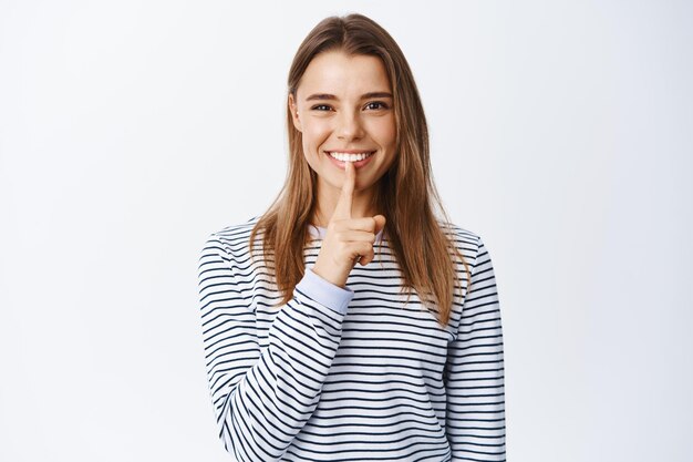 Image of pretty blond woman shushing and smiling, making shh taboo gesture and looking, asking to keep quiet, sharing a secret, standing on white