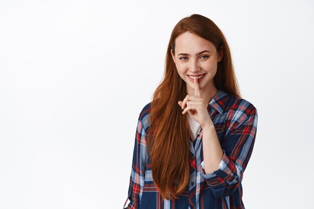 Image of pretty caucasian girl with red hair smiles, make shush, press finger to lips, quiet or taboo gesture, hushing with mysterious face, standing over white background.