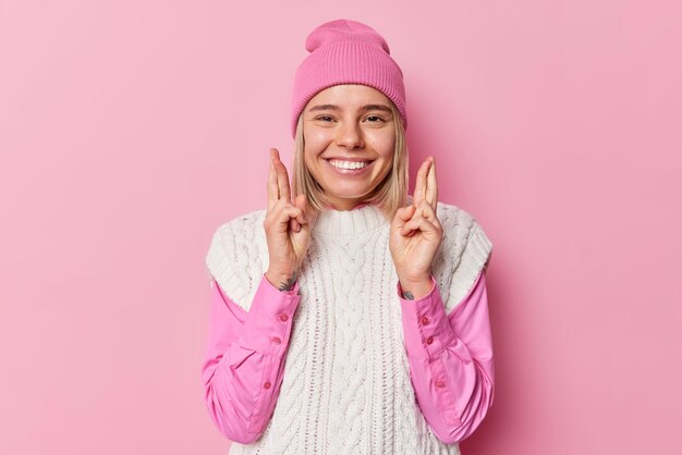 Image of pretty European girl aniticipates good positive news keeps fingers crossed smiles gladfully wears hat shirt and knitted white vest poses against pink background May my dreams come true