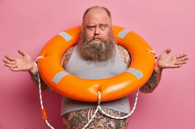 Image of puzzled overweight man spreads hands, poses with inflated lifebuoy, wears undersized t shirt, tattooed fat belly sticking out from it, going to swim in sea, isolated on pink wall