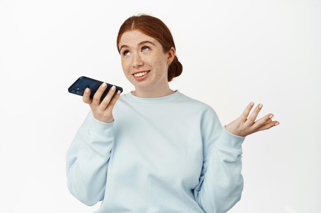 赤毛の白人の女の子が音声メッセージを録音し、スピーカーフォンで話し、モバイルアプリでジェスチャーとチャットをし、リラックスした笑顔の白い背景の画像。
