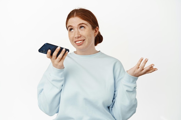 빨간 머리 백인 소녀의 이미지는 음성 메시지를 녹음하고, 스피커폰으로 말하고, 제스처를 취하고, 모바일 앱에서 채팅하고, 편안하고 흰색 배경에 웃고 있습니다.
