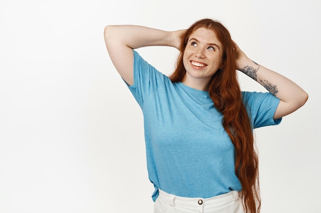 Foto gratuita immagine di una ragazza rossa sorridente rilassata sdraiata con le mani dietro la testa, che riposa e guarda in alto con una faccia felice e sognante, godendosi il fine settimana, il tempo libero, in piedi su sfondo bianco.