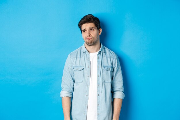 Image of reluctant and sad guy in casual outfit looking left, frowning and feeling upset, standing against blue background