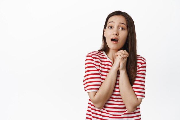 Image of romantic cute girl clench hands, looking with compassion and admiration, touched with smth beautiful, standing over white background in red t-shirt.