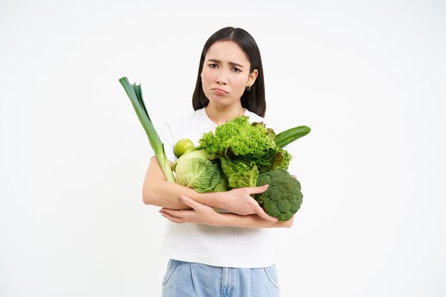 野菜を保持し、ダイエットの白い背景の上にすねている悲しいアジアの女の子の画像