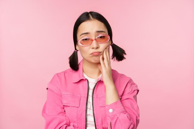 Изображение грустной азиатской девушки, дующейся, касающейся ее щеки, надутой разочарованной зубной боли, стоящей на розовом фоне