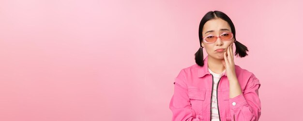 Image of sad asian girl sulking touching her cheek pouting disappointed has toothache standing over pink background