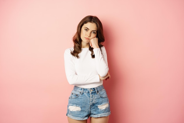 Free photo image of sad and bored, reluctant young woman looking at smth boring, feeling lonely, leaning head on fist with indifferent face expression, standing over pink background.