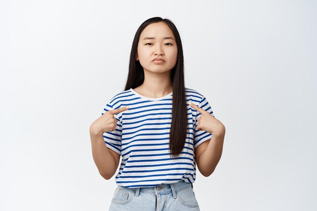 Image of sad brunette asian girl pointing fingers at herself making sulking upset face expression standing in striped tshirt over white background