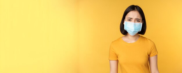 Image of sad and disappointed asian woman in medical mask frowning and looking displeased standing a