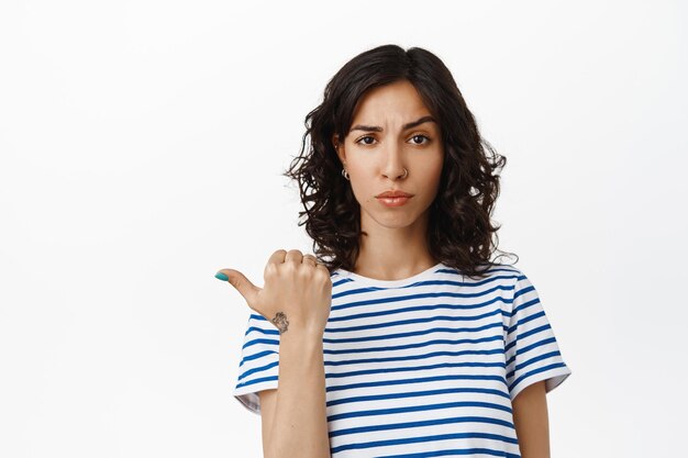Image of sad frowning girl pointing left, looking disappointed, complain on unfair thing, judging something bad awful, standing against white background