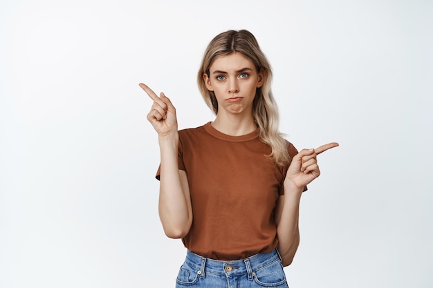 Image of sad young woman pointing fingers sideways at two lost options making upset face expression looking with regret standing against white background