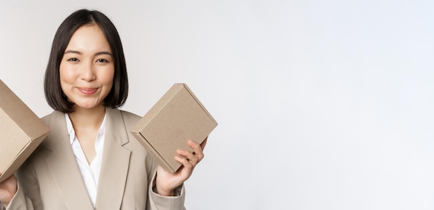 Изображение продавщицы азиатской бизнес-леди, держащей коробки с фирменным продуктом компании, улыбающейся в камеру, стоящей на белом фоне